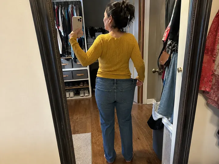 Dana G. with her back to a full-length mirror in her closet, holding the waistband of her jeans out from her hip.